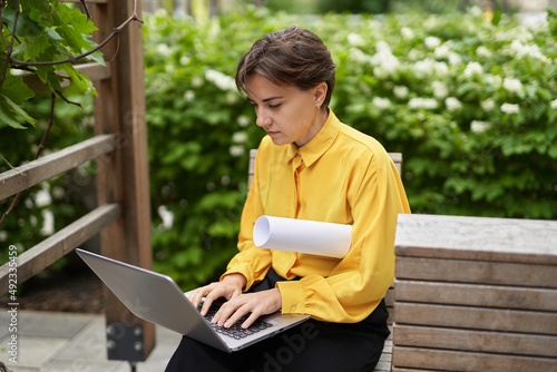 Business, finance industry or real estate concept. Successful smiling business lady in yellow blouse sitting on bench outdoor in park using laptop working or having online meeting. High quality image