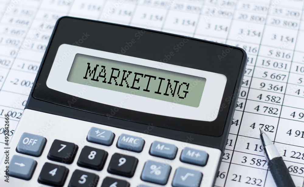 text MARKETING on the calculator screen on the background of reports on the table text MARKETING on the calculator screen on the background of reports on the table