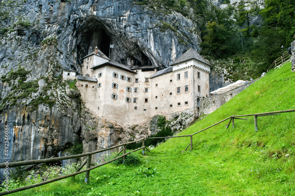 Predjama Castle in Slovenia. Old castle in the rock. Green meadow and behind it a large historic building with fortifications.