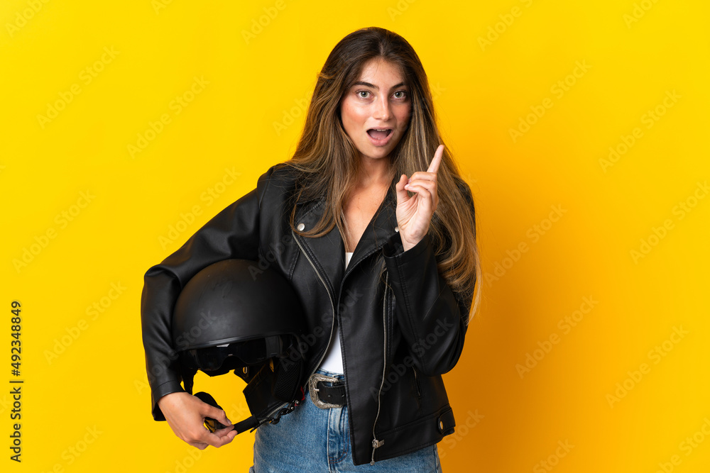Woman holding a motorcycle helmet isolated on yellow background intending to realizes the solution while lifting a finger up Woman holding a motorcycle helmet isolated on yellow background intending to realizes the solution while lifting a finger up