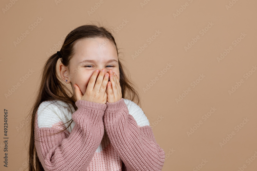 Portrait of young funny cute girl with long dark hair in white, pink sweater standing with closed eyes, blinking, covering mouth with hands on brown background. Silence, sneezing, laughing, emotion. Portrait of young funny cute girl with long dark hair in white, pink sweater standing with closed eyes, blinking, covering mouth with hands on brown background. Silence, sneezing, laughing, emotion.