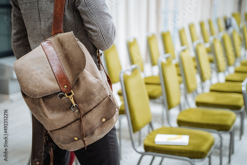 The backpack hangs on the shoulder of a man. Handmade leather bag in grey. A businessman stands with his back in a conference room.