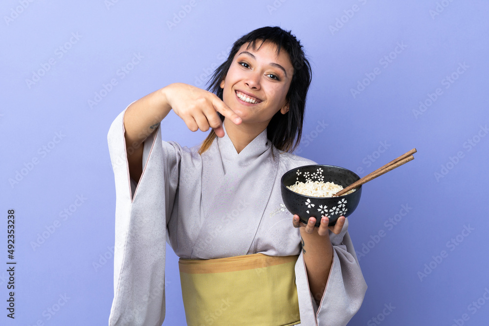Young woman wearing kimono over isolated blue background points finger at you with a confident expression while holding a bowl of noodles with chopsticks Young woman wearing kimono over isolated blue background points finger at you with a confident expression while holding a bowl of noodles with chopsticks