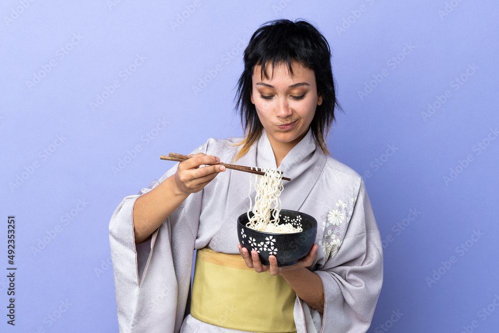 Young woman wearing kimono over isolated blue background holding a bowl of noodles with chopsticks