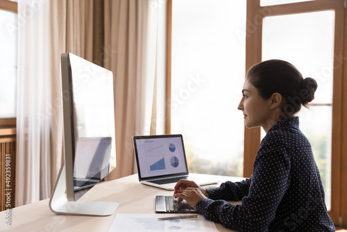 Fotografie Confident young Indian woman remote working specialist sit at office desk analyze market segments using statistic data graphs diagrams on pc