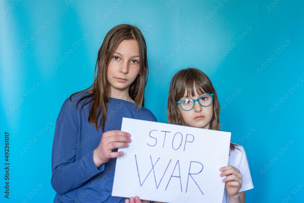 Portrait serious two school girls protesting war raises banner with inscription Stop War at blue background. Call to stop war, child against war, crisis in Ukraine. Portrait serious two school girls protesting war raises banner with inscription Stop War at blue background. Call to stop war, child against war, crisis in Ukraine.