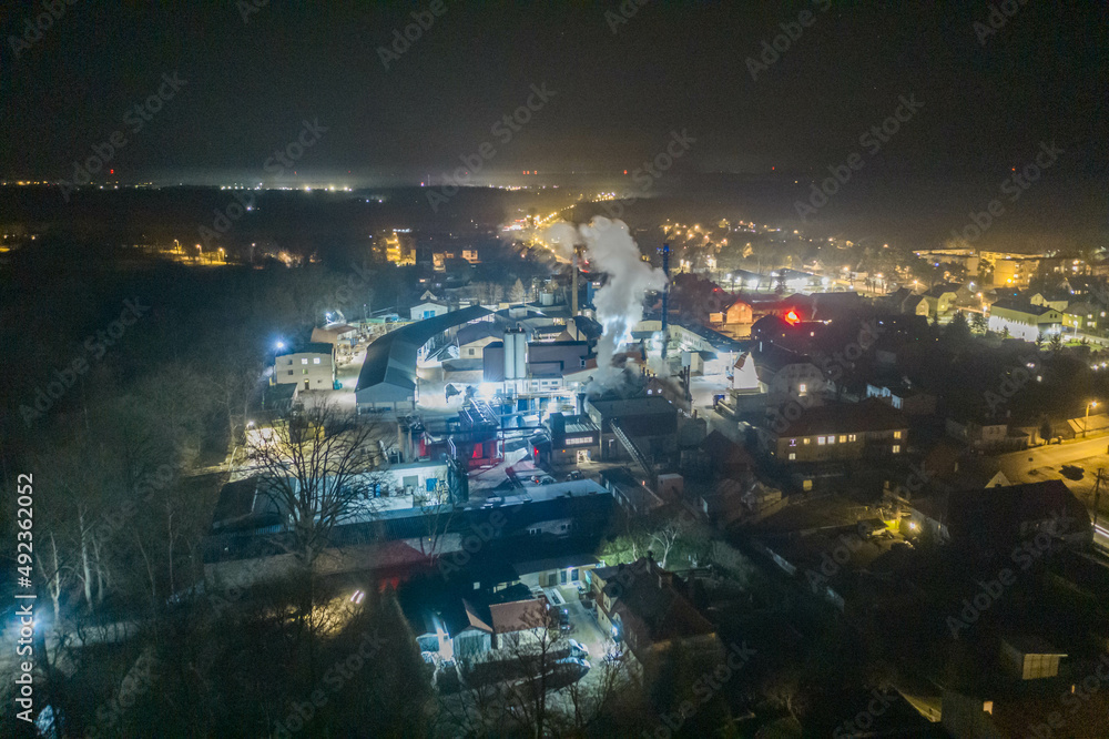 Panorama małego prowincjonalnego miasta w nocnej scenerii. W centrum kadru widać jaskrawo oświetloną fabrykę. Zdjęcie z wysokości wykonane z użyciem drona. - obrazy, fototapety, plakaty 