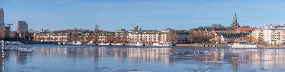 Panorama view at the icy bay Hammarby sjö with floes, apartment building and recreation area with commuting ferries a sunny winter day in Stockholm

