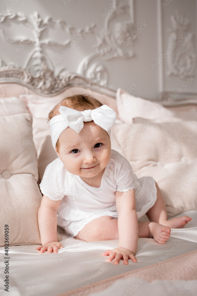 a beautiful baby girl in a white bodysuit and a headband is sitting on a white bed in a room with a beautiful interior a beautiful baby girl in a white bodysuit and a headband is sitting on a white bed in a room with a beautiful interior