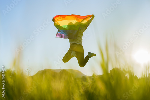 Unrecognizable man jumping with LGBT flag in a field at sunset