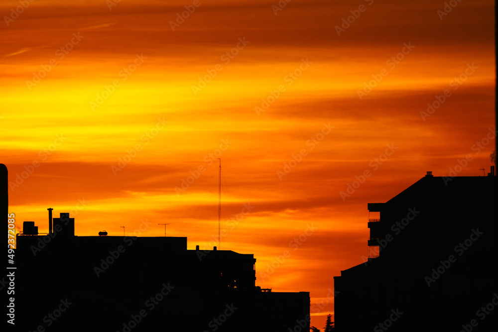 Fototapeta premium Dramatic cloudy sky at sunset in urban area. Silhouette buildings with the sun on yellow and orange background