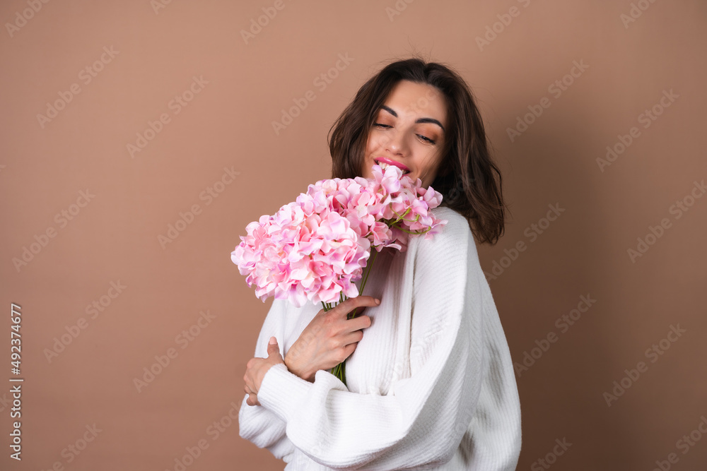 A young woman with wavy voluminous hair on a beige background with bright pink lipstick lip gloss in a white sweater holds a bouquet of pink flowers A young woman with wavy voluminous hair on a beige background with bright pink lipstick lip gloss in a white sweater holds a bouquet of pink flowers
