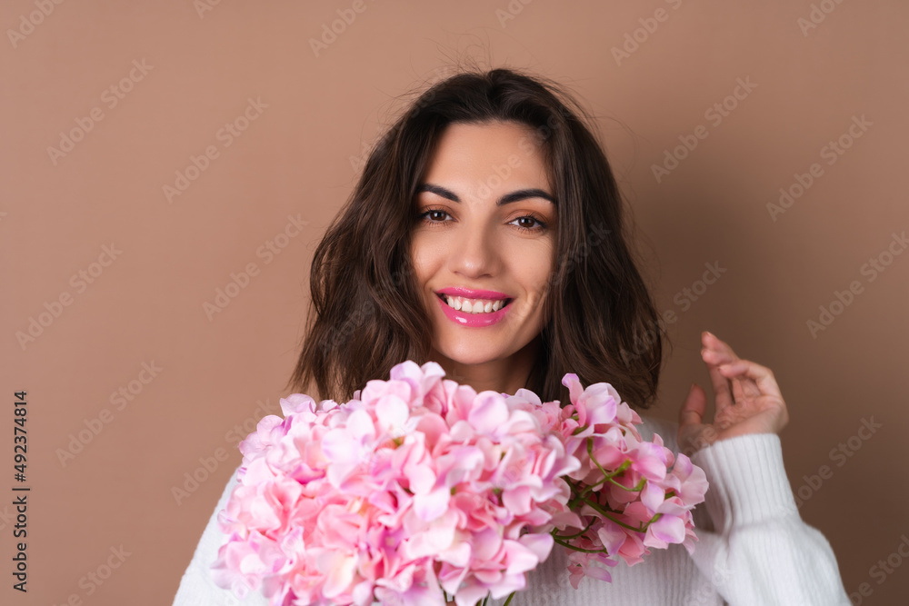 A young woman with wavy voluminous hair on a beige background with bright pink lipstick lip gloss in a white sweater holds a bouquet of pink flowers A young woman with wavy voluminous hair on a beige background with bright pink lipstick lip gloss in a white sweater holds a bouquet of pink flowers