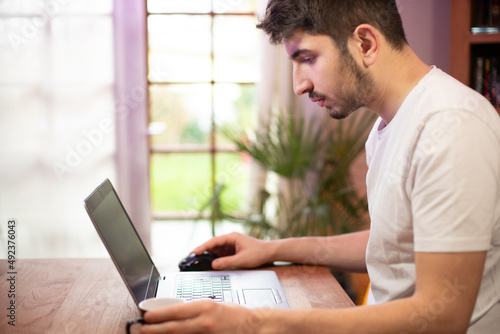 Jeune homme employé de bureau travaille devant son ordinateur portable en buvant un café. Il travaille à la maison, à distance en télé travail. On voit le jardin en arrière plan