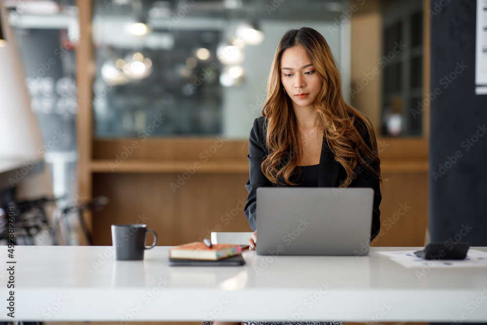 © David - Charming Asian businesswoman sitting in the office with a digital laptop computer. Excited Asian businesswoman raising hands to congratulate while working in a modern office,