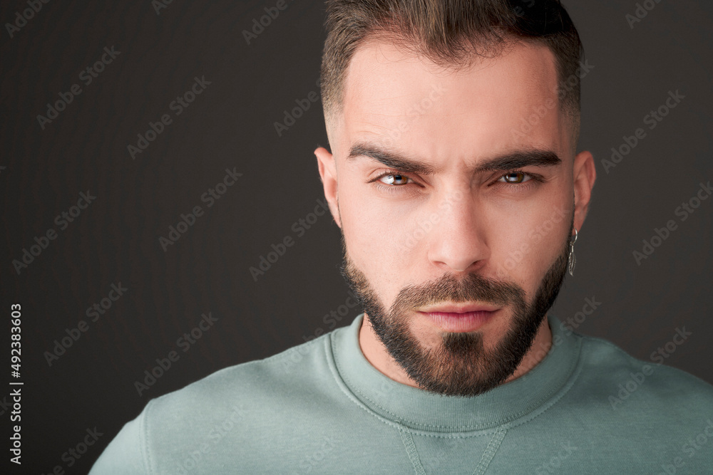 Male model shot in studio. Serious and confident expression. Angry or upset look and a casual style. Headshot of man with a beard, brown hair, brown eyes on a grey solid background. Male model shot in studio. Serious and confident expression. Angry or upset look and a casual style. Headshot of man with a beard, brown hair, brown eyes on a grey solid background.