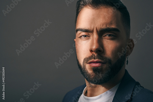 Male model shot in studio. Serious and confident expression. Angry or upset look and a casual style. Headshot of man with a beard, brown hair, brown eyes on a grey solid background. 