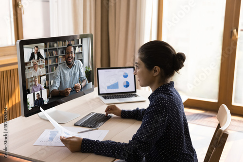 Young Indian lady confer with diverse teammates on distance discuss marketing report using conference app on desktop computer. Woman sales expert meet colleagues at online remote briefing by videocall