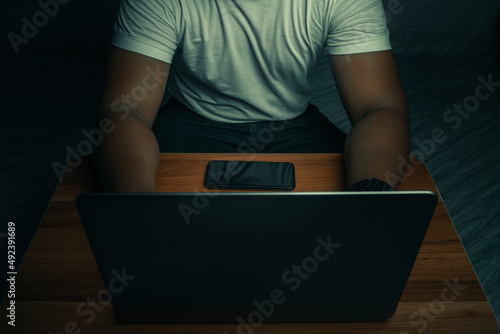 A man in a white shirt sits on a laptop in a dark room. with light shining down. internet concept addiction.topview.
