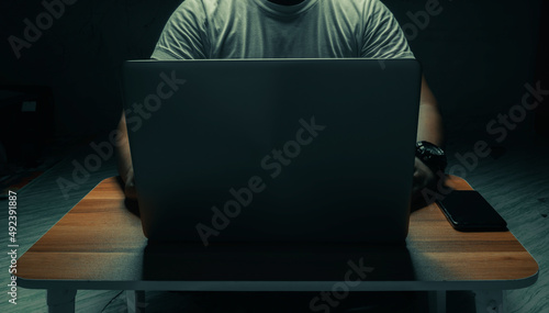 A man in a white shirt sits on a laptop in a dark room. with light shining down. internet concept addiction.topview.