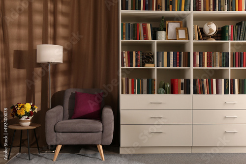 Cozy home library interior with comfortable armchair, floor lamp and collection of books on shelves