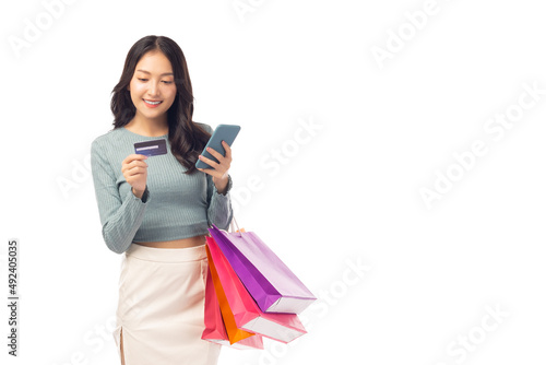 Beautiful Asian woman carrying shopping bags with using credit card and mobile phone standing over isolated on white background Smiling young girl purchasing online through phone using credit card