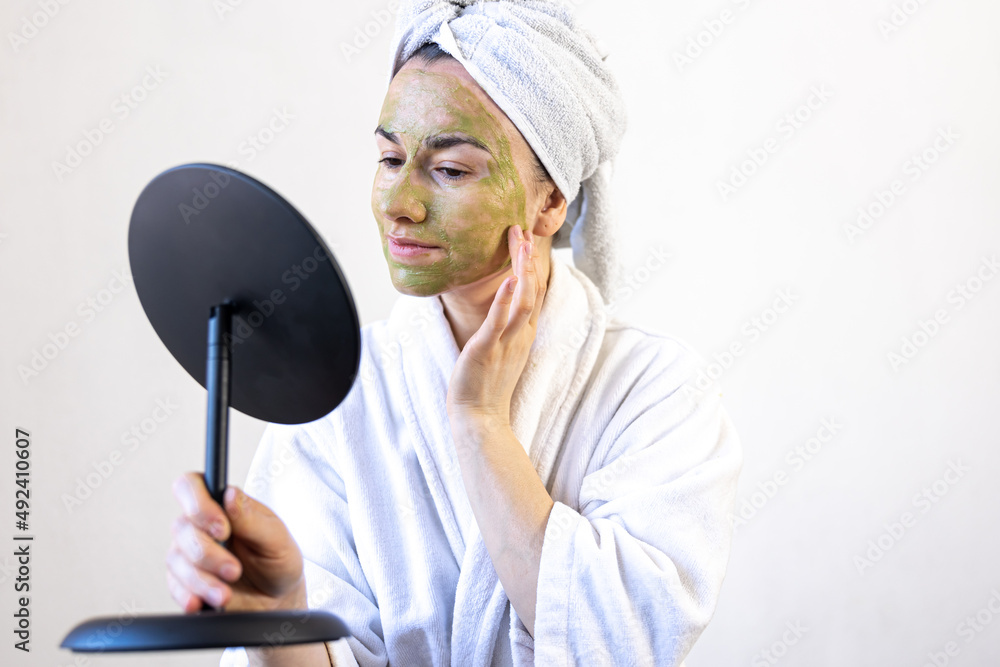 Young woman with a green mask on her face in a bathrobe in front of a mirror. Young woman with a green mask on her face in a bathrobe in front of a mirror.