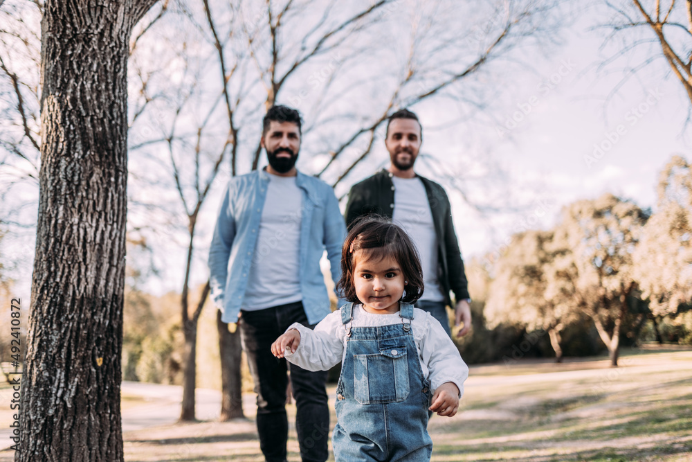 A little girl walking through the park followed by her parents. Gay family concept.