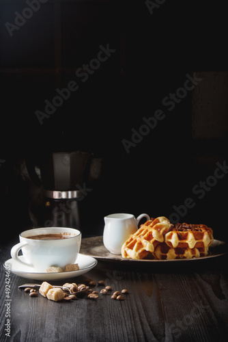 Homemade belgian waffles, white ceramic cup of coffee, milk, teaspoon and coffee beans. Dark rustic background.