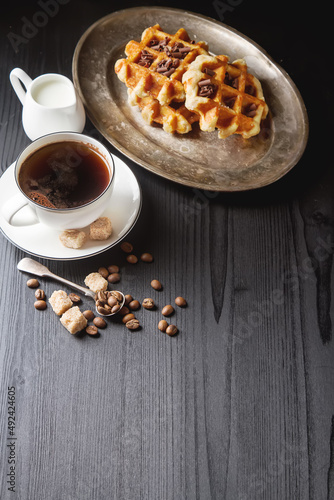 Homemade belgian waffles, white ceramic cup of coffee, milk, teaspoon and coffee beans. Dark rustic background. Space for text in the center.