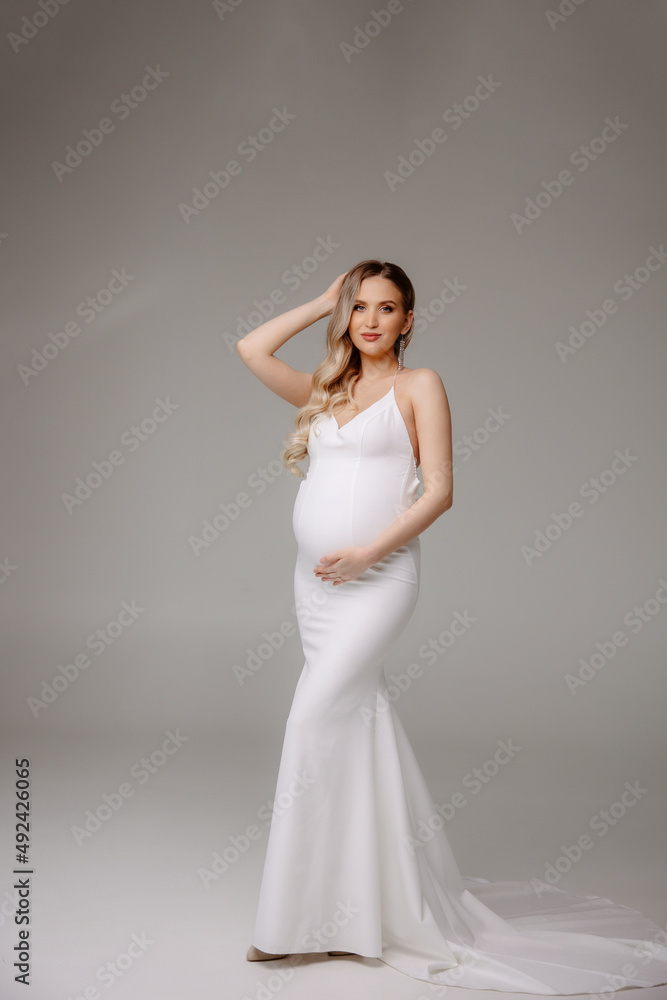 Happy pregnant woman in white long mermaide silhouette dress gently hugs touch belly with hands, studio background. Beautiful pregnancy, motherhood, expecting baby, family concept. Free space for text Happy pregnant woman in white long mermaide silhouette dress gently hugs touch belly with hands, studio background. Beautiful pregnancy, motherhood, expecting baby, family concept. Free space for text