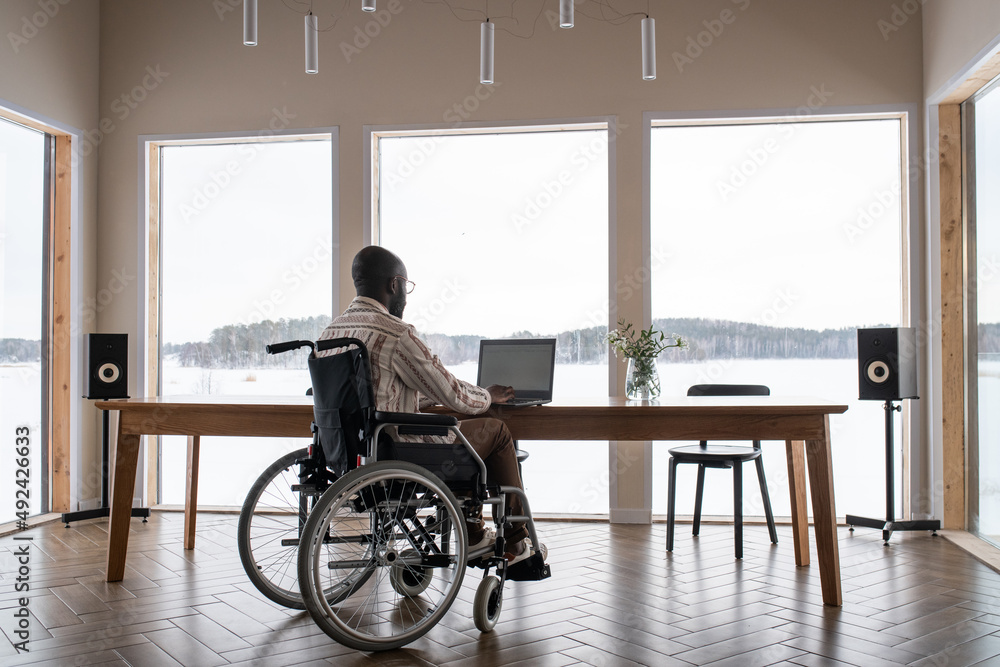 African American man with disability working in front of laptop while sitting by table in front of lapge window in living room African American man with disability working in front of laptop while sitting by table in front of lapge window in living room