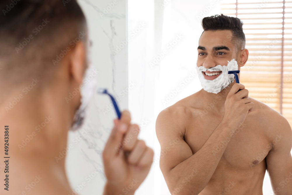 Handsome Shirtless Arab Guy Shaving Face While Looking At Mirror In Bathroom