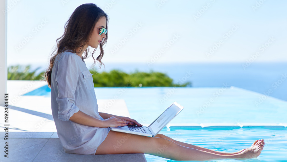 Browsing by the poolside. Shot of a young woman using her laptop by the poolside.