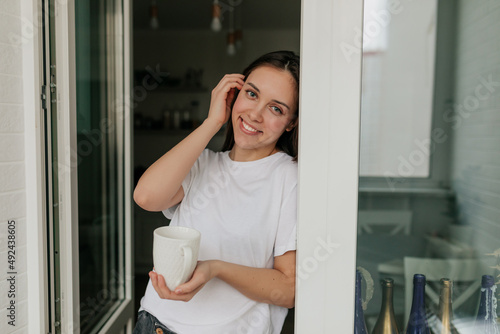 Adorable lovely woman smiling and touching her hair while posing at camera with morning cup with coffee at home in the kitchen
