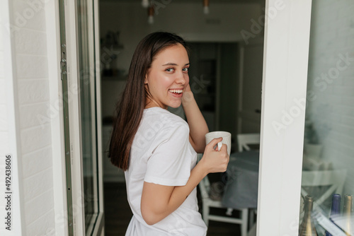 Pretty happy woman with dark hair in white t-shirt is holding cup with coffee and looking at camera with smile while having breakfast at home Free Download Pretty happy woman with dark hair in white t-shirt is holding cup with coffee and looking at camera with smile while having breakfast at home - GFXLoad