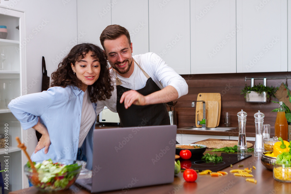 Happy couple using laptop computer preparing healthy food diet vegetable salad at home together. Woman and man are searching recipes, ordering shopping online, watching cooking class in kitchen.