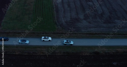 a column of six sports cars snake around the track in the middle of the field on the road at night. In the evening, a number of cars drive along the road, changing places. drone view photo