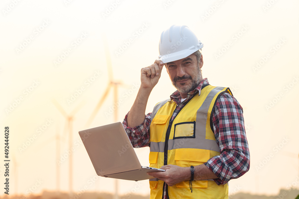Portrait of power engineer wearing safety jacket and hardhat with laptop computer working at outdoor field site that have wind turbine at the background. Portrait of power engineer wearing safety jacket and hardhat with laptop computer working at outdoor field site that have wind turbine at the background.