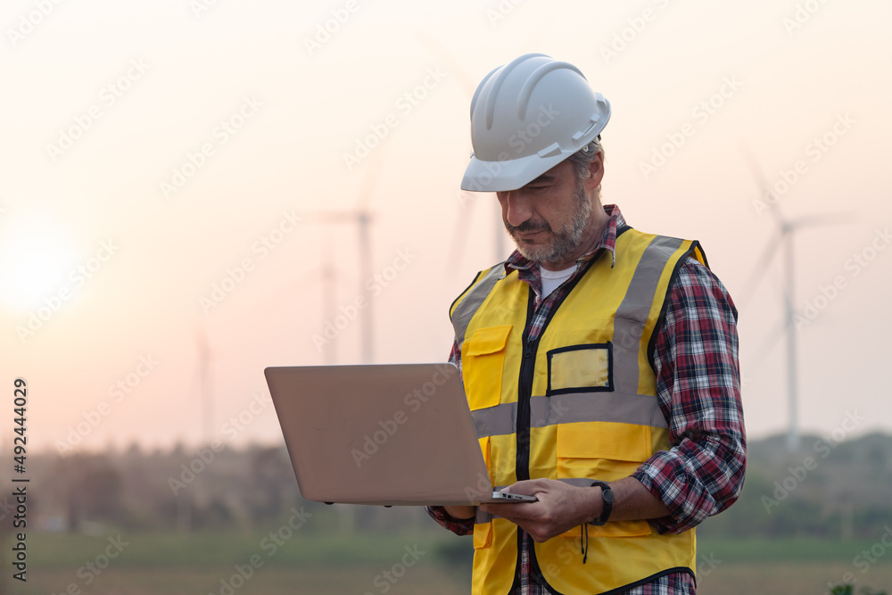Portrait of power engineer wearing safety jacket and hardhat with laptop computer working at outdoor field site that have wind turbine at the background.