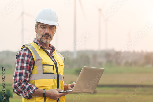 Portrait of power engineer wearing safety jacket and hardhat with laptop computer working at outdoor field site that have wind turbine at the background.