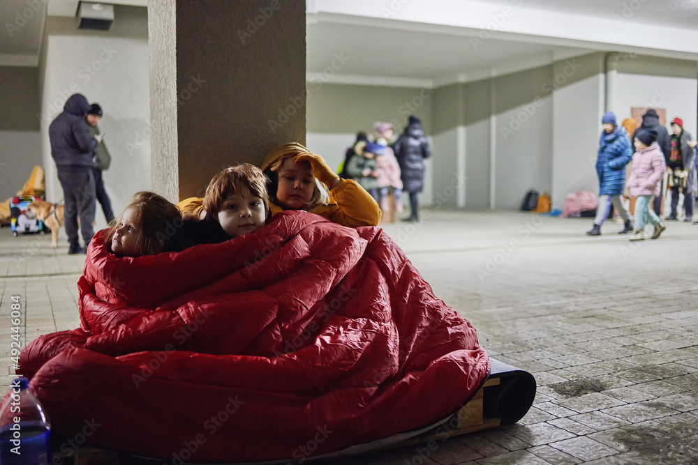 group of little kids, siblings sitting quietly on wooden pallet on the floor in bomb proof shelter during air-raid attack. Sharing one blanket during freezing night. russian invasion to Ukraine - obrazy, fototapety, plakaty 