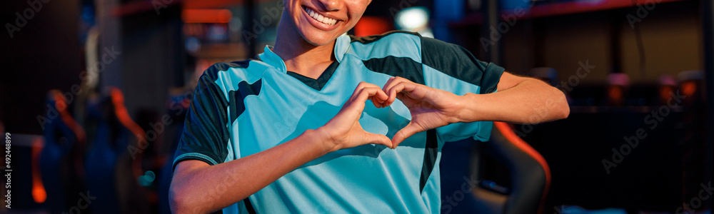Young African woman esports gamer in blue t-shirt showing heart sign by hands with happy facial expression in cyber club