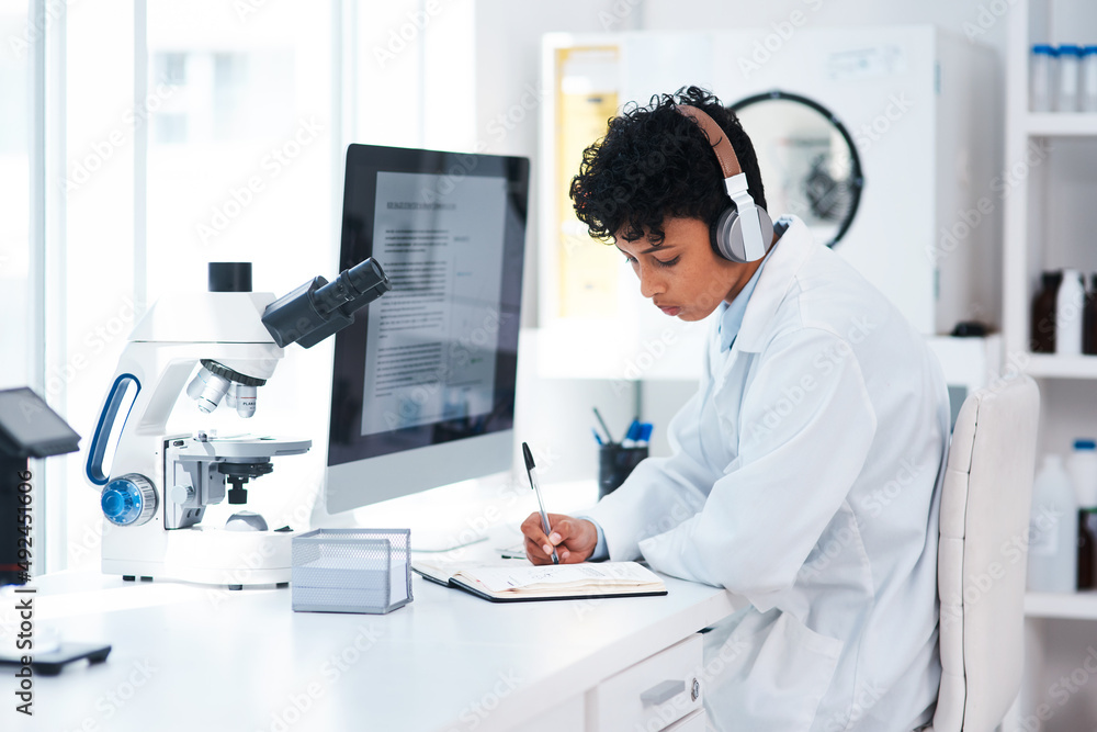 Shes one productive pathologist. Shot of a young scientist wearing headphones while writing notes and working on a computer in a lab. Shes one productive pathologist. Shot of a young scientist wearing headphones while writing notes and working on a computer in a lab.
