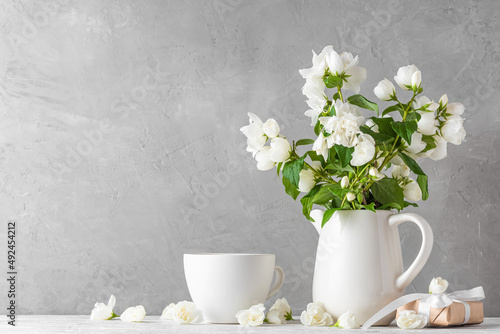 White jasmine blossom flowersbouquet in a vase with gift box and coffee cup on white wooden table. Still life photo