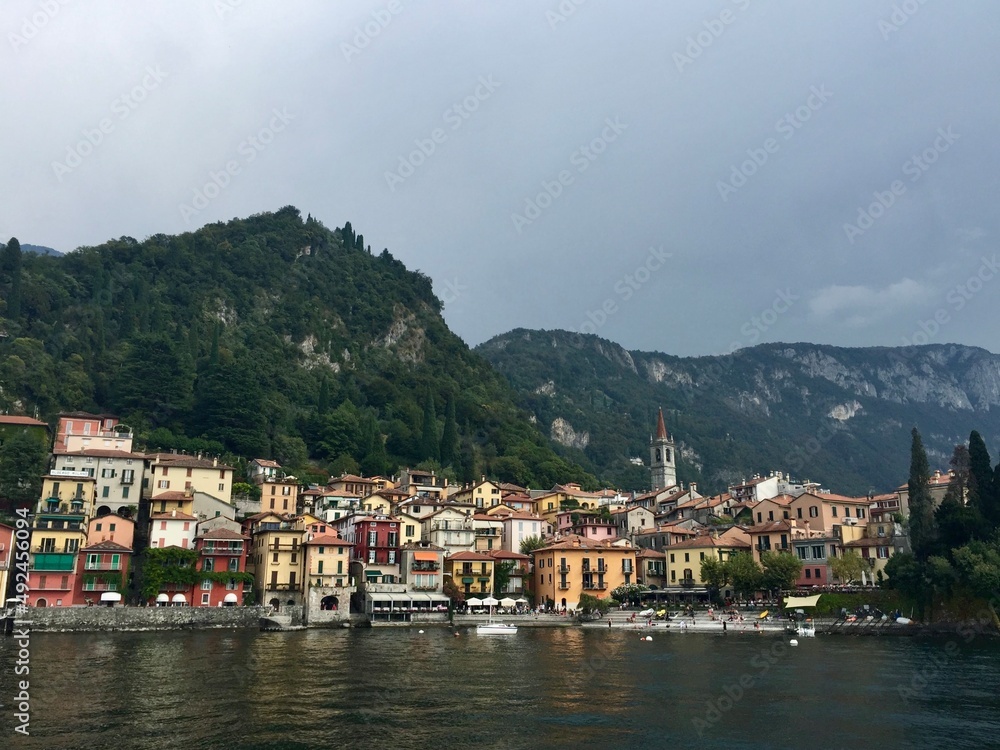 Varenna view, Italy. Colourful coastal houses view from Como lake. Hills and mountains. Forest, park, cloudy day, yellow buildings, bell tower.