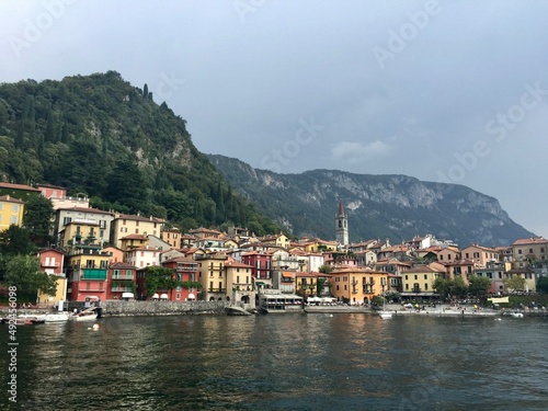 Varenna view, Italy. Colourful coastal houses view from Como lake. Hills and mountains. Forest, park, cloudy day, yellow buildings, bell tower. © Alla