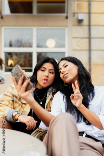 Two Latin female friends taking a selfie gesturing with their smartphone. Latin sisters having good times together in the street.