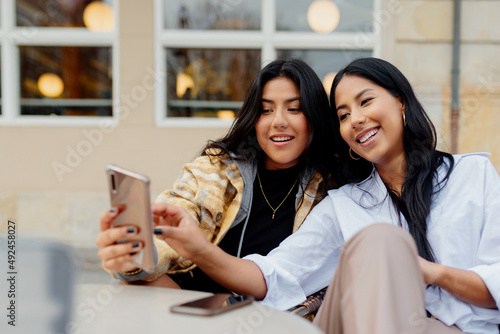 two hispanic sisters taking a picture with their smart phone in the street. Young latin girls using a mobile phone and having fun happily.