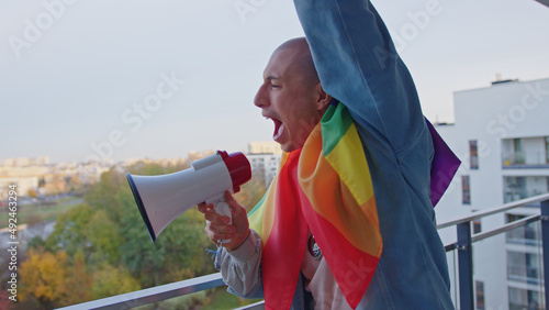caucasian proud bisexual man fighting for equality and same sex marriage, wrapped in rainbow flag screams aside in megaphone from his balcony. High quality photo